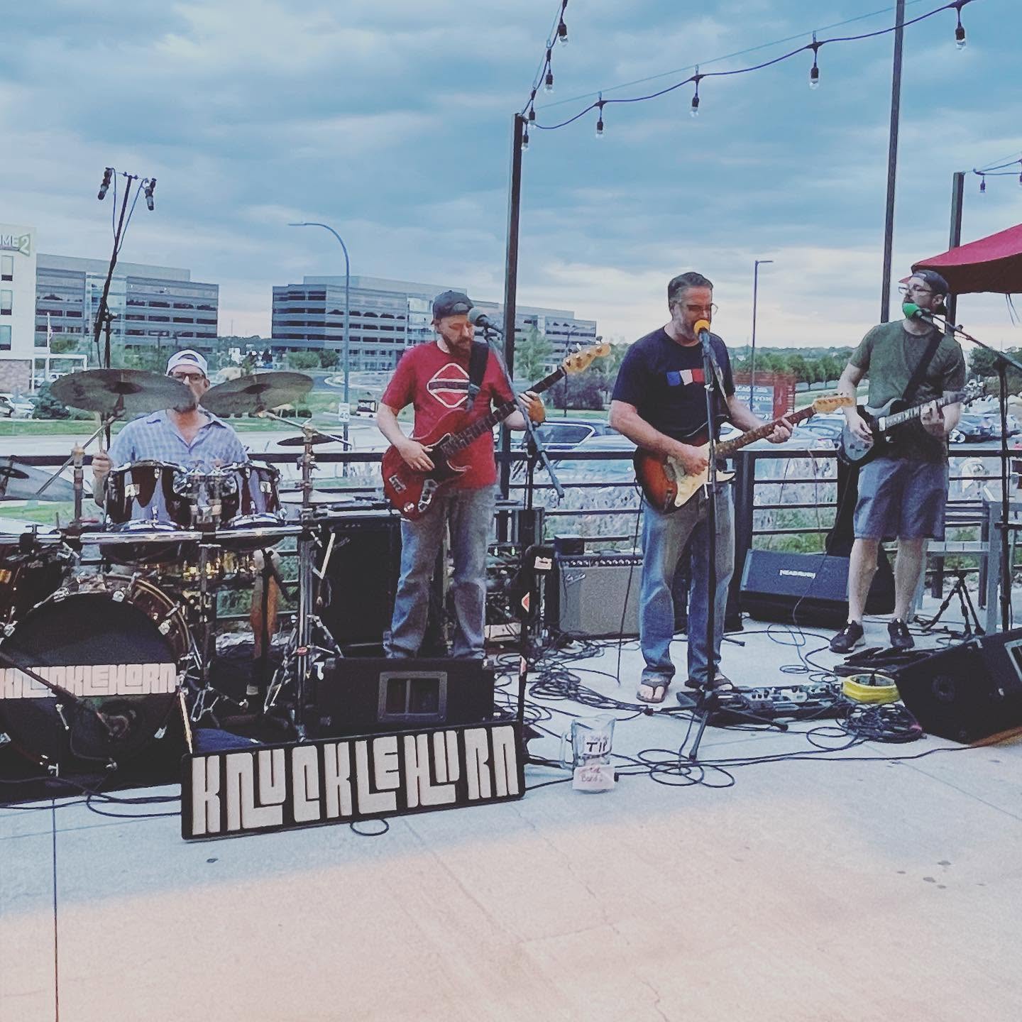 Knucklehorn playing an outdoor brewery patio in daylight, with the band banner in front of the drum kit